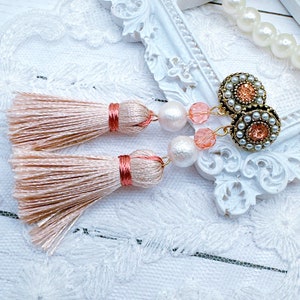 May include: A pair of blush-colored tassel earrings with gold-tone accents. The earrings feature a cluster of pearls and peach-colored beads. The tassels are made of a silky material and are tied with a matching thread.