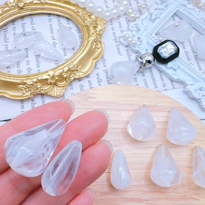 May include: A collection of seven white teardrop-shaped glass beads with a cloudy, translucent appearance. The beads are arranged on a wooden surface.