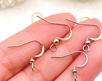 Puede incluir: Primer plano de varios ganchos para pendientes en tonos dorados y plateados. Los ganchos tienen un diseño curvo con una pequeña bola y un lazo en la parte inferior. Los ganchos están diseñados para la fabricación de joyas.