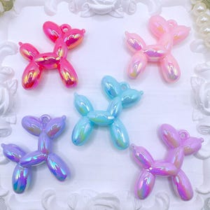 May include: Five iridescent balloon dog charms in pink, light pink, blue, purple, and lavender. Each charm has a loop for attaching to jewelry or other crafts. The charms are arranged on a white, decorative surface.