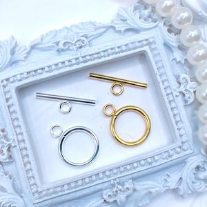 May include: Close-up of jewelry findings, including silver and gold toggle clasps and rings. The clasps have a bar and ring design, perfect for necklaces and bracelets. The findings are displayed in a white decorative frame, with a pearl necklace in the background.