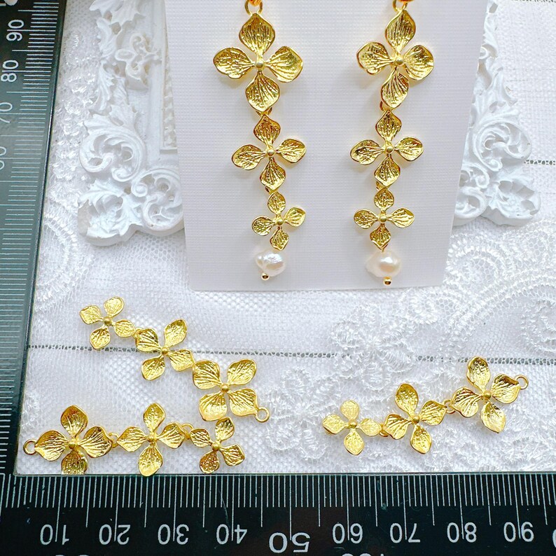 May include: Gold-tone flower-shaped earrings with pearl accents. The earrings are shown on a white surface with a ruler in the background. There are also several smaller flower-shaped charms on the surface.
