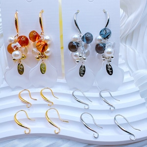 May include: Two pairs of earrings with a white flower-shaped charm. One pair has amber and white beads, the other has gray and white beads. Both pairs have silver hooks.