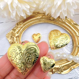May include: Four gold heart-shaped locket pendants with intricate designs. The lockets are on a white surface with white flowers and a gold frame.