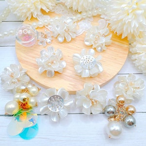 May include: A collection of white floral embellishments and pearl accents. The assortment includes various flower designs, some with pearl centers and others with metallic details. Several pieces feature dangling pearl and sequin clusters.