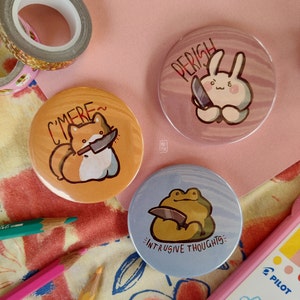 May include: Three round pins with colorful backgrounds and cartoon characters holding knives. The pins say "C'mere", "Perish", and "Intrusive Thoughts".