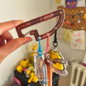 Könnte beinhalten: Ein klarer Acryl-Schlüsselanhänger mit einem braunen, messerförmigen Karabinerhaken und drei kleineren Schlüsselanhängern. Die Schlüsselanhänger zeigen einen gelben Frosch, eine Cartoonfigur und ein farbenfrohes Design. Die Website-Adresse mitsujuice.etsy.com ist sichtbar.