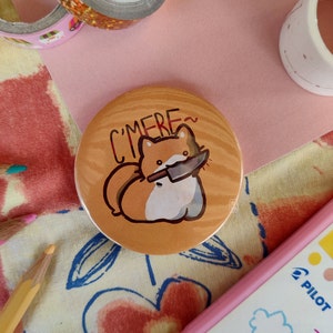 以下が含まれることがあります： ナイフを口にくわえた茶色と白の動物の漫画イラストが描かれた丸いボタン。動物の上には「C'MERE~」の文字。ボタンは木目調の背景です。