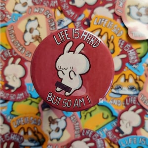 May include: A red button with a white bunny illustration and the text "Life is hard but so am I". The bunny is holding a black bar in front of it.