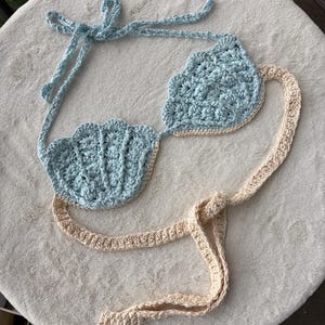 May include: A crochet bikini top featuring two light blue shell-shaped cups with scalloped edges. The top includes light blue neck ties and a long beige waist tie that connects the base of the cups, all crafted with a textured yarn stitch pattern.