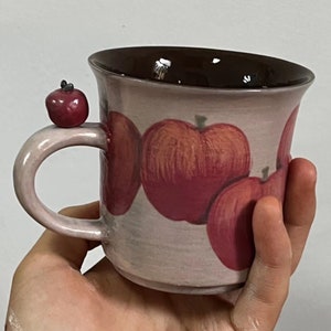 Könnte beinhalten: Eine Keramiktasse mit braunem Rand und einem Henkel in Form eines Apfels. Die Tasse ist mit einem Design aus rosa und roten Äpfeln verziert.