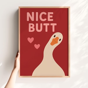以下が含まれることがあります： 赤い背景にピンク色の文字で「NICE BUTT」と書かれた額入りのアートプリント。ベージュの体、オレンジ色のくちばし、2つのピンクのハートを持つ漫画のガチョウが描かれています。