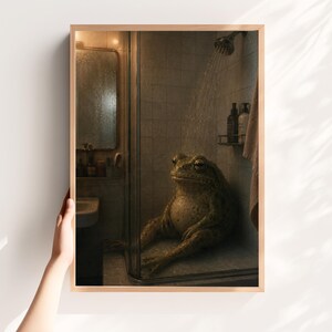 May include: A framed print depicts a large frog sitting in a shower stall. The frog is green and brown, with a contemplative expression. The showerhead is on, and water streams down. The background includes a mirror, toiletries, and a towel.