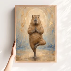May include: A framed wall art print featuring a brown capybara standing on one leg in a yoga pose with hands pressed together. The background is an abstract painting with gold, beige, and soft blue brushstrokes forming a circular halo effect.