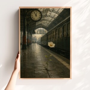 May include: A framed wall art print depicting a single white flower growing through a crack in a dark, wet train station platform. A vintage train car sits on the tracks beside a large clock hanging from the arched ceiling of the station.