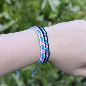 May include: Three friendship bracelets in the image. One bracelet is black, another is white, pink, and blue, and the third is blue and pink. The bracelets are tied on the wrist with a green background.