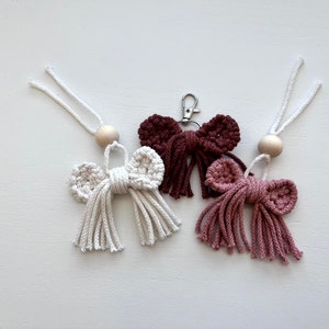 May include: Three crocheted bow keychains with wooden beads and tassels. The bows are in shades of brown, pink, and white.