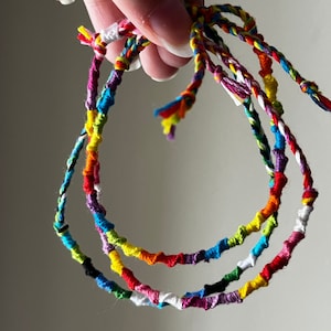 May include: Three colorful friendship bracelets made of woven string in shades of red, yellow, blue, green, purple, white, and black.