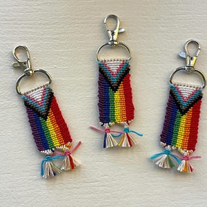 May include: Three handmade keychains with rainbow-colored woven designs and silver-tone clasps. Each keychain features a colorful pattern with a triangle design at the top and small tassels at the bottom. The keychains are displayed on a light-colored surface.