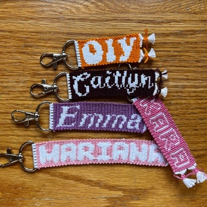 May include: Four colorful woven keychains with white lettering. The keychains spell out the names "Olivia", "Faithlyn", "Emma", and "Mariah".