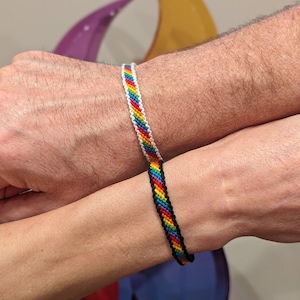 May include: Two friendship bracelets with a rainbow pattern. One bracelet is white with a rainbow stripe, and the other is black with a rainbow stripe.