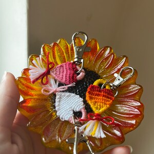 May include: Three crocheted heart-shaped keychains with tassels. The keychains are in pink, white, and orange and red. The keychains are on a yellow sunflower-shaped dish.