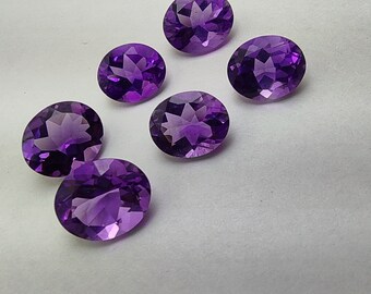 Könnte beinhalten: Sechs ovale geschliffene Amethyst-Edelsteine, jeder mit einem violetten Farbton. Die Edelsteine sind in einem Halbkreis auf einer weißen Oberfläche angeordnet.