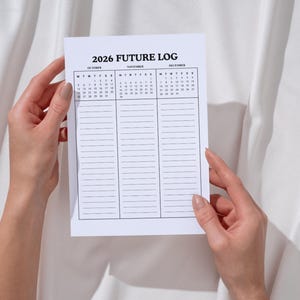 May include: A white paper planner titled "2026 Future Log" is held by two hands. The planner features a calendar for October, November, and December, along with lined sections for notes. The background is a white fabric.