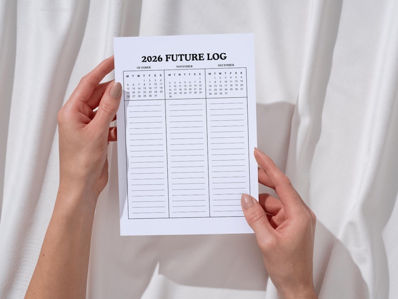 May include: A white paper planner titled "2026 Future Log" is held by two hands. The planner features a calendar for October, November, and December, along with lined sections for notes. The background is a white fabric.