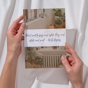 May include: A printed card with a quote by Will Rogers: "Don't wait to buy real estate. Buy real estate and wait." The card features images of a window with plants and a radiator. The card is held against a white fabric background.