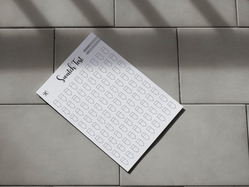 May include: A white swatch test sheet with a grid of small, outlined shapes, labeled "Swatch Test" in cursive script. The number "90" is in the lower left corner. The sheet is on a gray tiled surface.