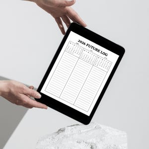 May include: A black tablet displaying a 2026 Future Log calendar with lined sections. The tablet is held by hands against a white background. The calendar layout includes months and dates.