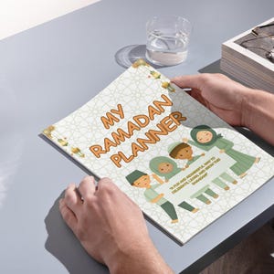 May include: A white and green planner with the text "My Ramadan Planner" in orange. The planner features a colorful illustration of children holding a sign that says "It's fun and meaningful way to celebrate, learn and reflect this Ramadan."