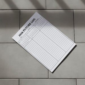 May include: A white 2026 Future Log planner with a calendar and lined sections for notes. The planner is angled on a gray tiled surface, with shadows across the page.