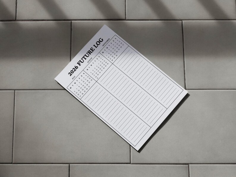 May include: A white 2026 Future Log planner with a calendar and lined sections for notes. The planner is angled on a gray tiled surface, with shadows across the page.