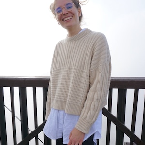 Puede incluir: Una mujer lleva un suéter de punto beige con trenzas y una camisa blanca abotonada debajo. Está sonriendo y lleva gafas de color azul. El fondo es una imagen borrosa de una barandilla de madera y niebla.