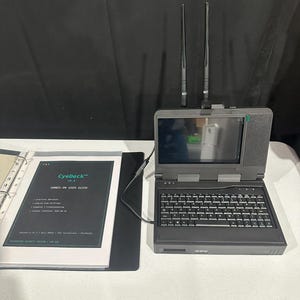 May include: A dark gray portable computer device with a built-in screen, keyboard, and two vertical antennas. Next to it is a binder with a cover labeled CyeDeck v0.4 HANDS-ON USER GUIDE, Raspberry Pi 4, Kali ARM64, and 7-inch Touchscreen.