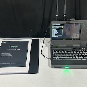 May include: A dark gray portable computer with a built-in keyboard, screen, and two antennas, displaying a green command line interface. Beside it lies a black binder containing a CyeDeck v0.4 hands-on user guide with white text on a dark background.