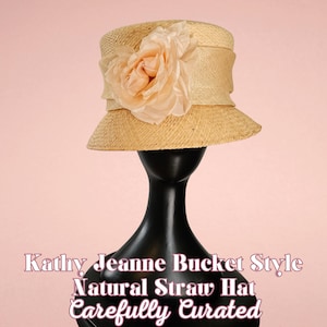 May include: A natural straw bucket hat with a wide brim and a peach-colored fabric flower. The hat is displayed on a black mannequin head against a pink background. Text on the image reads "Kathy Jeanne Bucket Style Natural Straw Hat Carefully Curated."