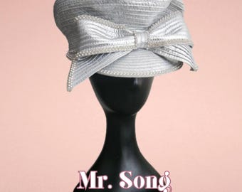 May include: A silver hat with a structured crown and a wide, pleated bow detail on the front. The edges of the bow and the brim are accented with small white pearls. The text Mr. Song Carefully Curated is visible at the bottom of the image.