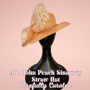 May include: A peach-colored straw hat with a wide brim and a large bow adorned with white lace floral embroidery. The hat is displayed on a black mannequin head against a pink background. Text on the image reads "Mr. John Peach Sinamay Straw Hat Carefully Curated."