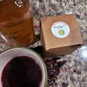 May include: A brown paper box with the text "Warm Wishes Blend Hibiscus, Clove, and cinnamon" and a colorful logo. The box is sitting on a table with a glass jar of honey and a cup of red tea.