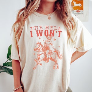 May include: A cream colored t-shirt with the text "THE HELL I WON'T" in pink. A vintage illustration of a woman riding a horse is printed on the shirt.