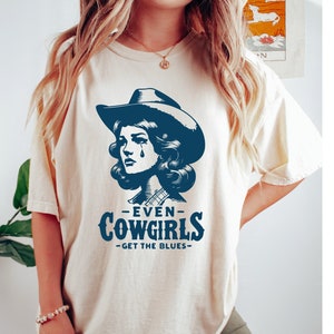 May include: A cream colored t-shirt with a blue graphic of a woman wearing a cowboy hat and the text "Even Cowgirls Get The Blues".