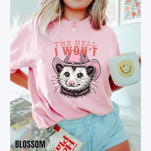 Pu&ograve; includere: Maglietta rosa con un'immagine di un opossum che indossa un cappello da cowboy rosa e il testo "The Hell I Won't".