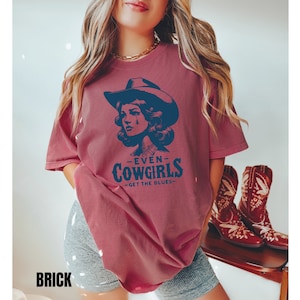 May include: A maroon t-shirt with a distressed look featuring a vintage illustration of a woman wearing a cowboy hat and the text "Even Cowgirls Get the Blues".