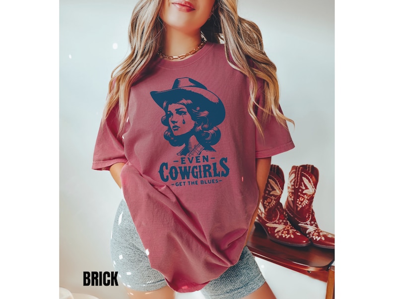 May include: A maroon t-shirt with a distressed look featuring a vintage illustration of a woman wearing a cowboy hat and the text "Even Cowgirls Get the Blues".