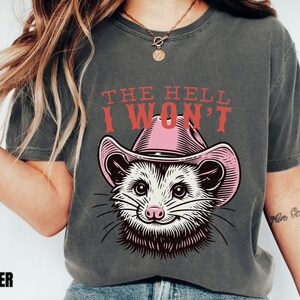 Pu&ograve; includere: Una maglietta grigia con una grafica di un opossum che indossa un cappello da cowboy rosa. Il testo "THE HELL I WON'T" &egrave; stampato in rosso sopra l'opossum.