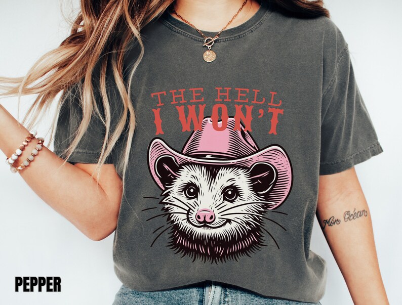 Pu&ograve; includere: Una maglietta grigia con una grafica di un opossum che indossa un cappello da cowboy rosa. Il testo "THE HELL I WON'T" &egrave; stampato in rosso sopra l'opossum.