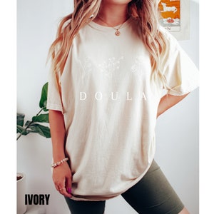 May include: A cream colored t-shirt with a white floral design and the word "DOULA" printed on the front. The t-shirt is being worn by a person with long blonde hair. The word "IVORY" is printed in black at the bottom of the image.